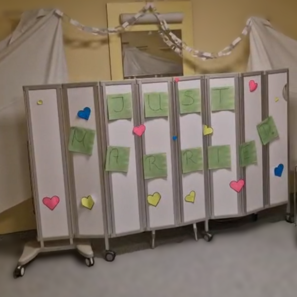 Auf einer Stellwand sind grüne Zettel mit Buchstaben, die das Wort Just Married ergeben, rundherum sind Herzen dekoriert. Im Hintergrund sieht man eine Girlande aus Ringen.